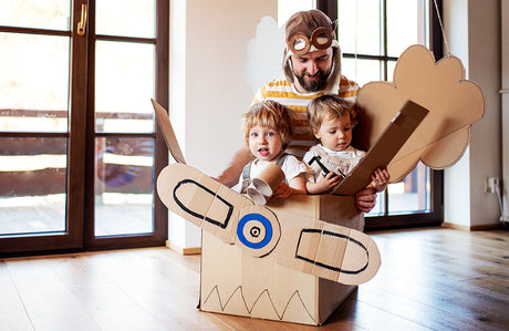 Ein Vater und zwei Kleinkinder spielen mit einem Kartonflugzeug zu Hause.