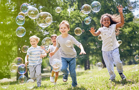 Kinder rennen auf einer Wiese und freuen sich über fliegende Seifenblasen.